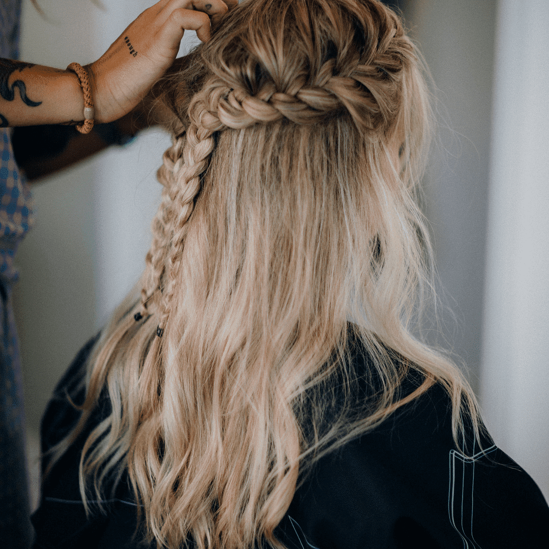 Hairstylist braiding blonde woman's hair in a salon. Tattoo and bracelet visible on stylist's arm.