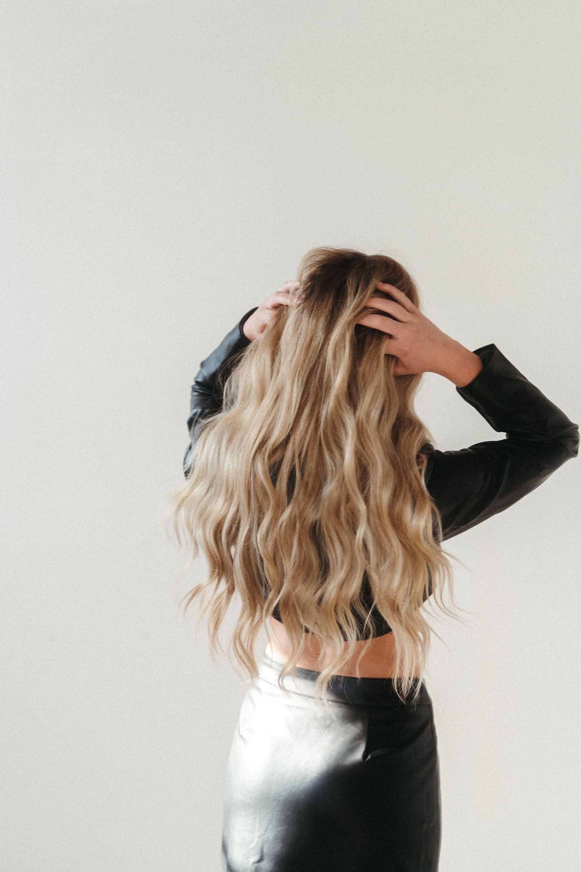 Woman with long wavy blonde hair in black outfit facing away, hands in hair.