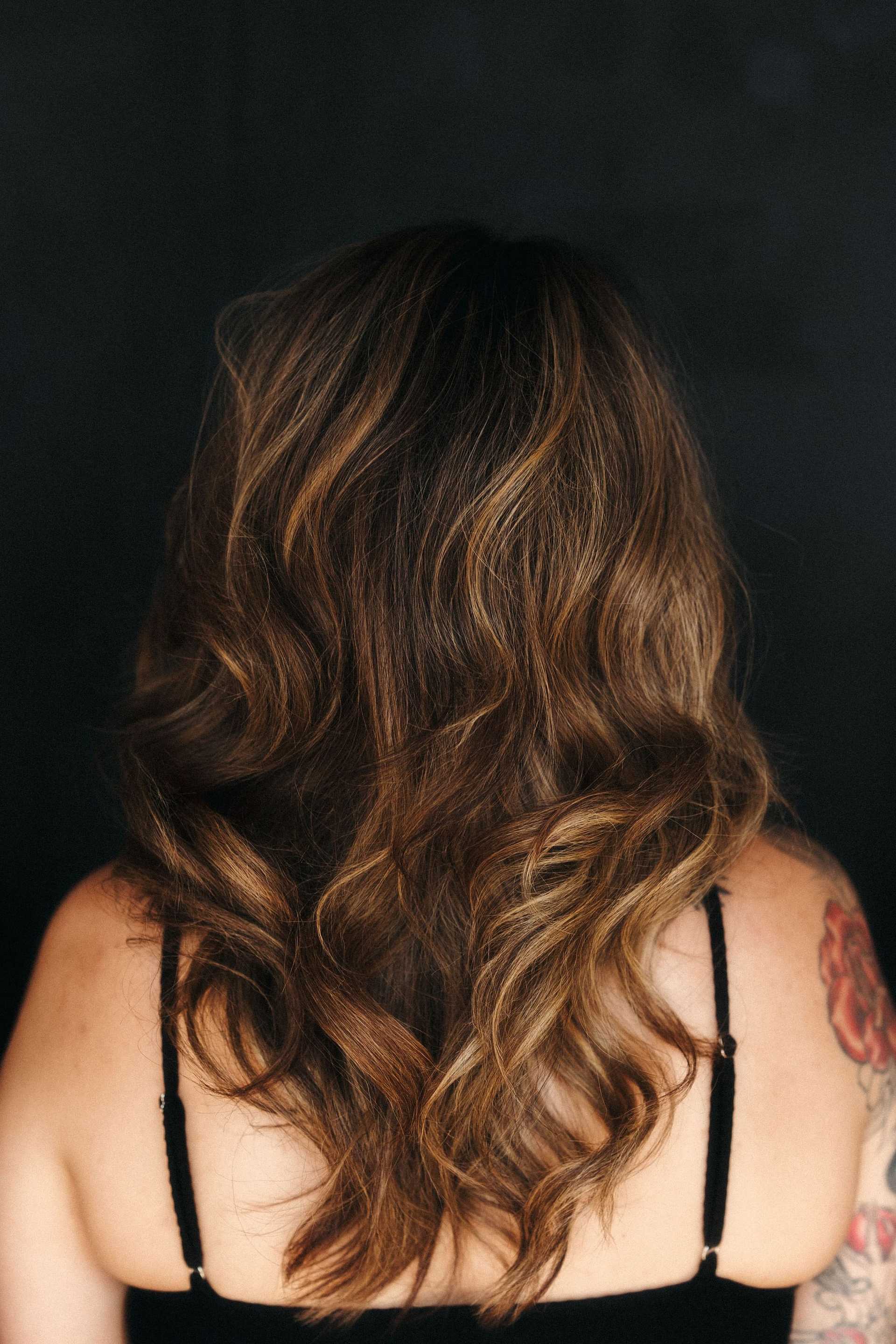 Back view of a woman with long, wavy brown hair against a dark background.